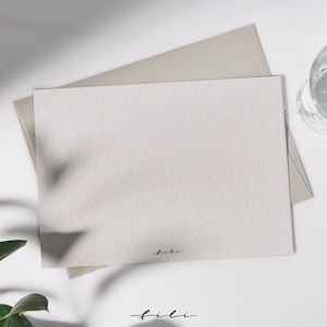 May include: A blank white card with a subtle textured surface. The card is sitting on top of a light brown envelope. The word "fili" is written in black ink at the bottom of the card.