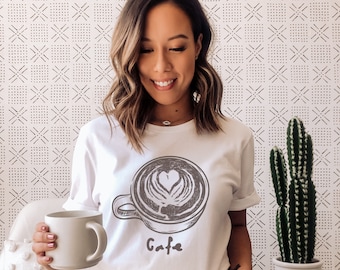 Puede incluir: Una camiseta blanca con una ilustración en blanco y negro de una taza de café con la palabra "Cafe" impresa debajo.