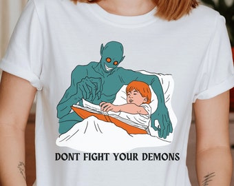 May include: White t-shirt with a graphic illustration of a child reading in bed with a teal demon. The text "DONT FIGHT YOUR DEMONS" is printed below the illustration. The shirt is made of a soft material.