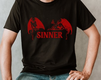 May include: Black t-shirt featuring a red graphic of two winged figures and the word "SINNER" in red block letters. The figures are in a stylized, illustrative style. The shirt is short-sleeved and made of a dark fabric.