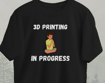 May include: Black t-shirt with white text that reads "3D Printing in Progress". A cartoon illustration of a person sitting in a yoga pose is above the text.