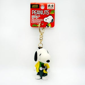 Pu&ograve; includere: Un portachiavi dorato con una statuina 3D di Snoopy vestito da samurai giapponese. Il portachiavi ha un'etichetta che recita "PEANUTS Costume Snoopy" in inglese e giapponese. L'etichetta riporta anche il testo "JAPAN collection" e "BUSHI" in inglese.