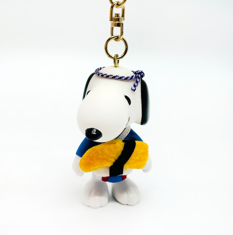 Pu&ograve; includere: Un cane di cartone animato bianco e nero che indossa un kimono blu e giallo che tiene un rotolo di sushi giallo e nero. Il cane indossa una fascia per capelli blu e bianca. Il cane &egrave; attaccato a un portachiavi dorato.