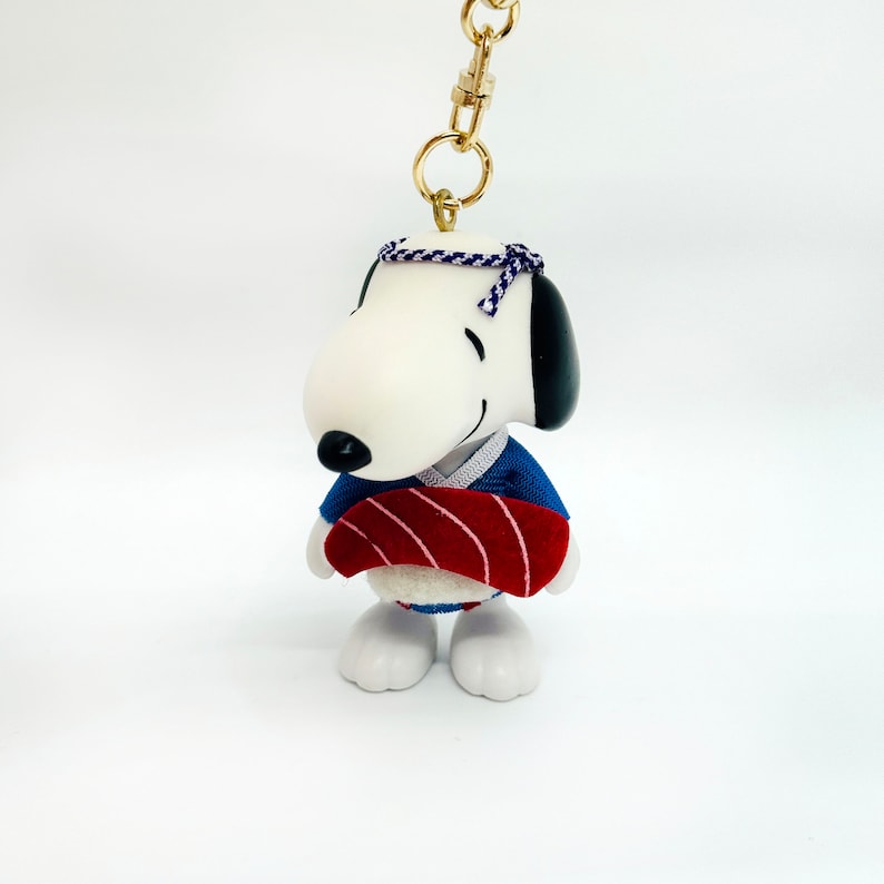 Pu&ograve; includere: Un cane di cartone animato bianco e nero che indossa un kimono blu e rosso che tiene un sushi rosso e bianco. Il cane indossa una fascia per capelli blu e bianca. Il cane &egrave; attaccato a una catena dorata.