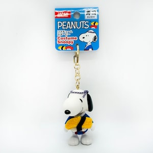 Pu&ograve; includere: Un portachiavi con Snoopy, un personaggio dei fumetti Peanuts, vestito con un costume da chef di sushi blu e giallo. Il portachiavi ha una catena dorata e un'etichetta blu e bianca con la scritta "Costume Snoopy" e "Peanuts".