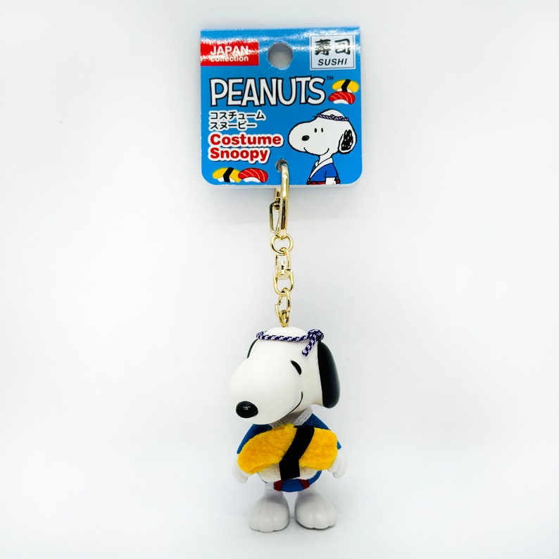 Pu&ograve; includere: Un portachiavi con Snoopy, un personaggio dei fumetti Peanuts, vestito con un costume da chef di sushi blu e giallo. Il portachiavi ha una catena dorata e un'etichetta blu e bianca con la scritta "Costume Snoopy" e "Peanuts".