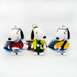 Pu&ograve; includere: Tre figure di Snoopy di peluche bianco che indossano abiti giapponesi tradizionali. Le figure indossano abiti blu, gialli e rossi con diversi accessori. Le figure sono su uno sfondo bianco.