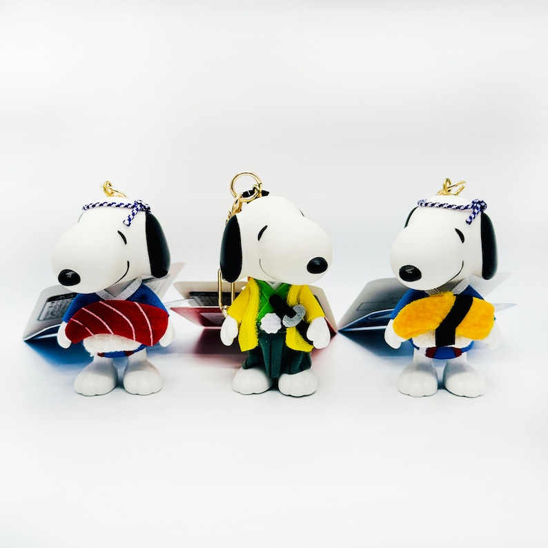 Pu&ograve; includere: Tre figure di Snoopy di peluche bianco che indossano abiti giapponesi tradizionali. Le figure indossano abiti blu, gialli e rossi con diversi accessori. Le figure sono su uno sfondo bianco.