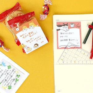 Puede incluir: Una composici&oacute;n de naturaleza muerta con art&iacute;culos de papeler&iacute;a y bocadillos japoneses. Una bolsa transparente de galletas con envoltorio rojo se encuentra junto a un cuaderno con una nota adhesiva rosa. Tambi&eacute;n hay un l&aacute;piz, un comp&aacute;s y una regla.