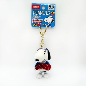 Pu&ograve; includere: Un portachiavi con Snoopy, un personaggio dei fumetti Peanuts, vestito con un costume da chef di sushi blu e rosso. Sta tenendo un rotolo di sushi rosso. Il portachiavi &egrave; attaccato a una catena dorata. La confezione &egrave; blu e bianca con il testo "PEANUTS Costume Snoopy" e "寿司 SUSHI".