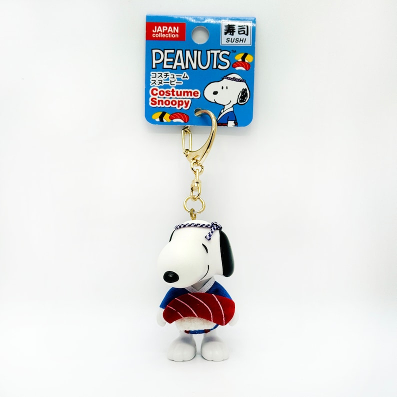 Pu&ograve; includere: Un portachiavi con Snoopy, un personaggio dei fumetti Peanuts, vestito con un costume da chef di sushi blu e rosso. Sta tenendo un rotolo di sushi rosso. Il portachiavi &egrave; attaccato a una catena dorata. La confezione &egrave; blu e bianca con il testo "PEANUTS Costume Snoopy" e "寿司 SUSHI".
