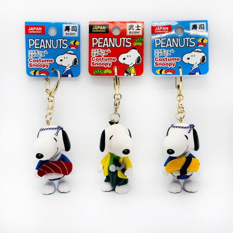 Pu&ograve; includere: Tre portachiavi con il personaggio di Snoopy dei Peanuts vestito con diversi costumi culturali giapponesi. I portachiavi sono etichettati "PEANUTS Costume Snoopy" e mostrano Snoopy vestito da chef di sushi, samurai e uomo giapponese tradizionale.