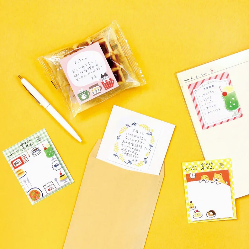 Puede incluir: Una colecci&oacute;n de art&iacute;culos de papeler&iacute;a sobre una superficie amarilla. Incluye un bol&iacute;grafo, notas adhesivas con texto japon&eacute;s e ilustraciones de comida y animales, un gofre y un sobre con una tarjeta.