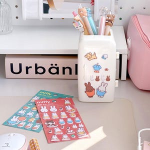 May include: A white mini refrigerator-shaped pencil holder with a variety of colorful stickers featuring a bunny character. The stickers are on the front of the holder and include a dog, a shirt, and a bunny wearing a blue shirt. Two packages of stickers featuring the same bunny character are on a white surface. The packages are labeled "Miffy" and feature a variety of colorful stickers.