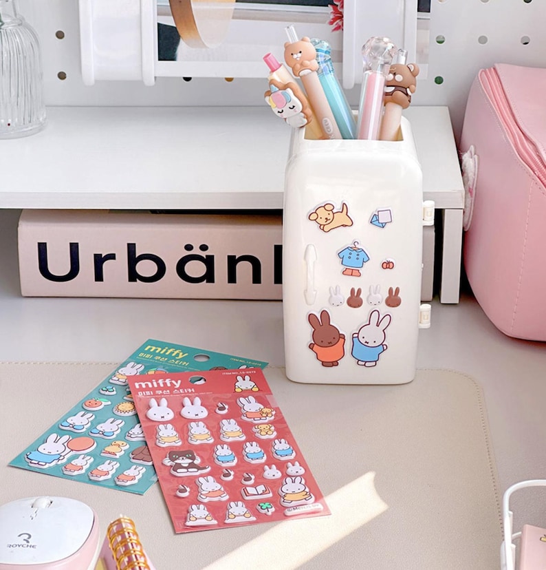 May include: A white mini refrigerator-shaped pencil holder with a variety of colorful stickers featuring a bunny character. The stickers are on the front of the holder and include a dog, a shirt, and a bunny wearing a blue shirt. Two packages of stickers featuring the same bunny character are on a white surface. The packages are labeled "Miffy" and feature a variety of colorful stickers.