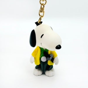Pu&ograve; includere: Una statuetta di Snoopy bianca e nera che indossa un kimono giallo e verde. La statuetta &egrave; attaccata a un portachiavi dorato.