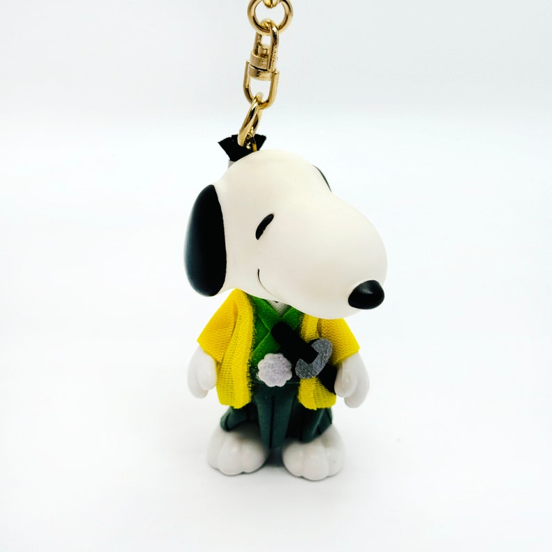 Pu&ograve; includere: Una statuetta di Snoopy bianca e nera che indossa un kimono giallo e verde. La statuetta &egrave; attaccata a un portachiavi dorato.