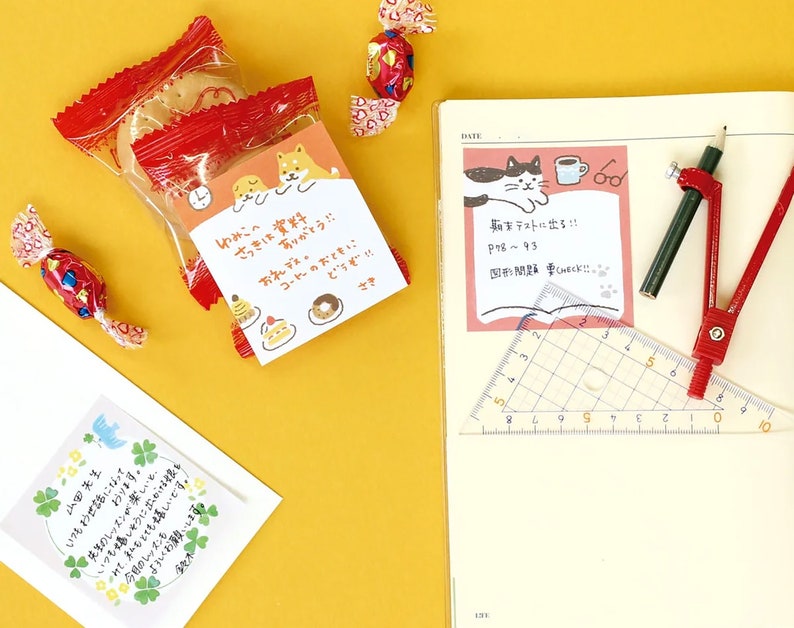 May include: A still-life composition featuring Japanese stationery and snacks. A clear plastic bag of cookies with a red wrapper sits next to a notebook with a pink sticky note. A pencil, compass, and ruler are also present. Candies and a card are scattered around.