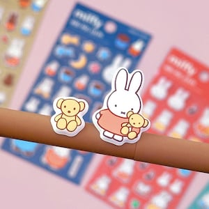May include: A sheet of stickers featuring a white bunny rabbit holding a teddy bear, a brown teddy bear, and other cute illustrations. The stickers are on a white background.