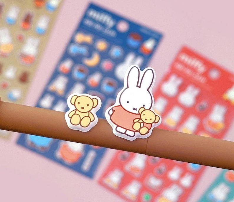 May include: A sheet of stickers featuring a white bunny rabbit holding a teddy bear, a brown teddy bear, and other cute illustrations. The stickers are on a white background.