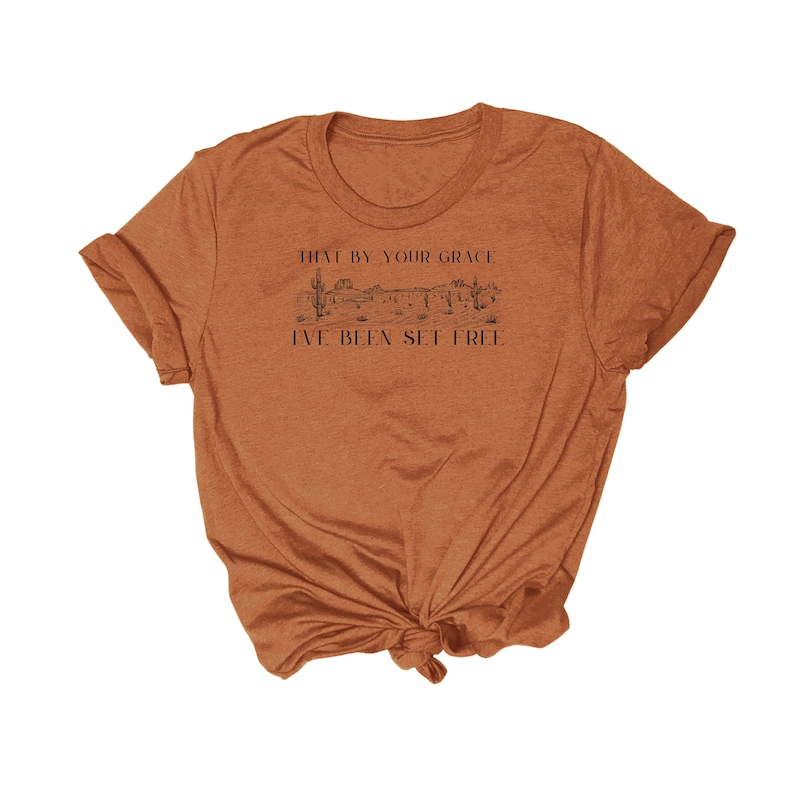 May include: A brown t-shirt with a graphic of a desert landscape and the text "That by your grace I've been set free".