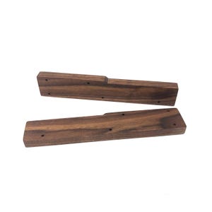 May include: Two wooden keyboard wrist rests. The rests are made of dark brown wood with a visible grain pattern. Each rest has a rectangular shape with a recessed area on one end and several small holes.