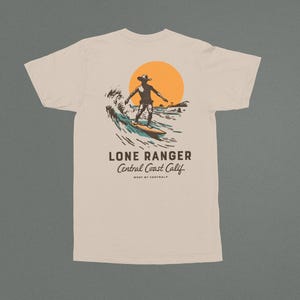 May include: A beige t-shirt with a graphic of a surfer riding a wave in front of a setting sun. The text "LONE RANGER Central Coast Calif. WEST BY CENTRAL" is printed below the graphic.