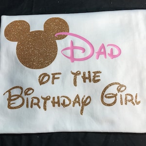 May include: White t-shirt with gold glitter Mickey Mouse ears and the text "Dad of the Birthday Girl" in gold glitter. The word "Dad" is in pink.