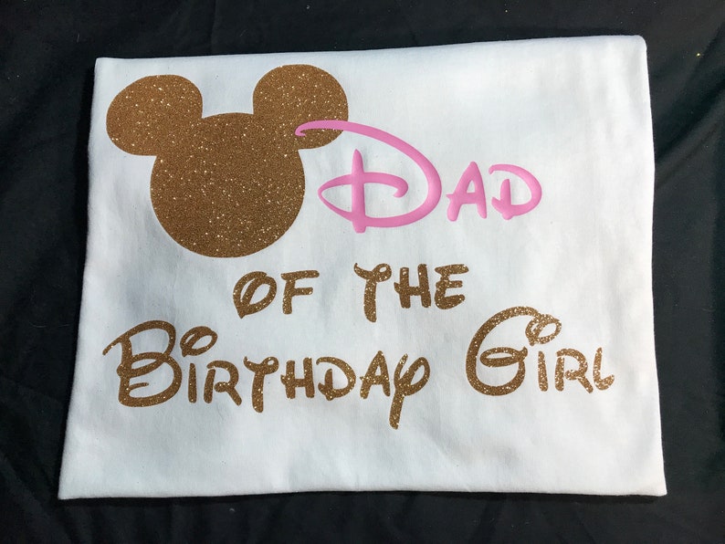 May include: White t-shirt with gold glitter Mickey Mouse ears and the text "Dad of the Birthday Girl" in gold glitter. The word "Dad" is in pink.