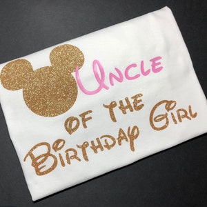 May include: White t-shirt with gold glitter Mickey Mouse ears and the text "Uncle of the Birthday Girl" in gold glitter lettering. The word "Uncle" is in pink glitter lettering.