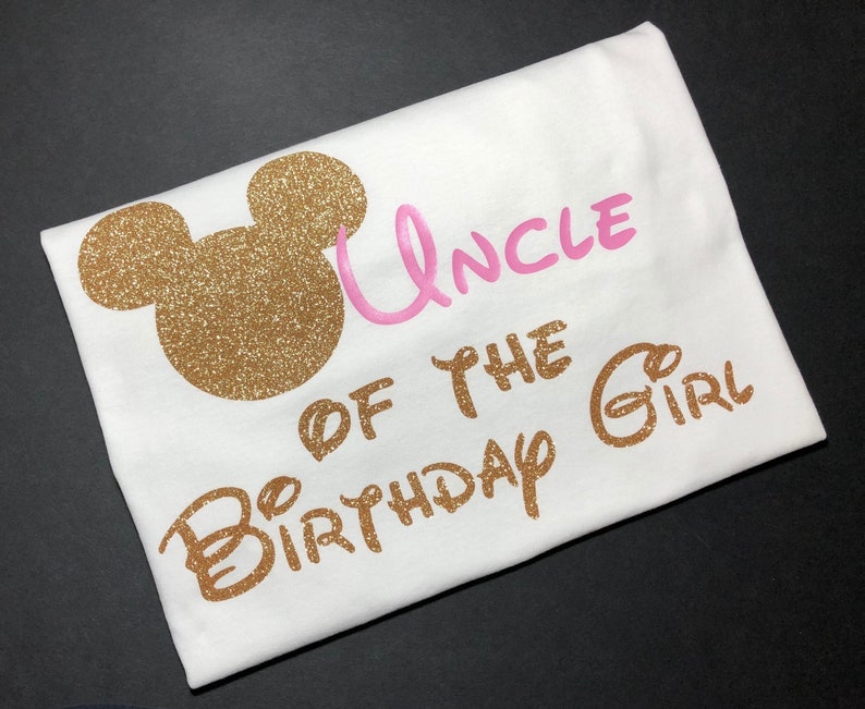May include: White t-shirt with gold glitter Mickey Mouse ears and the text "Uncle of the Birthday Girl" in gold glitter lettering. The word "Uncle" is in pink glitter lettering.