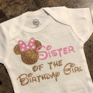 May include: White baby onesie with gold glitter lettering that says "Big Sister of the Birthday Girl". The onesie has a pink and gold glitter Minnie Mouse head on the left side.