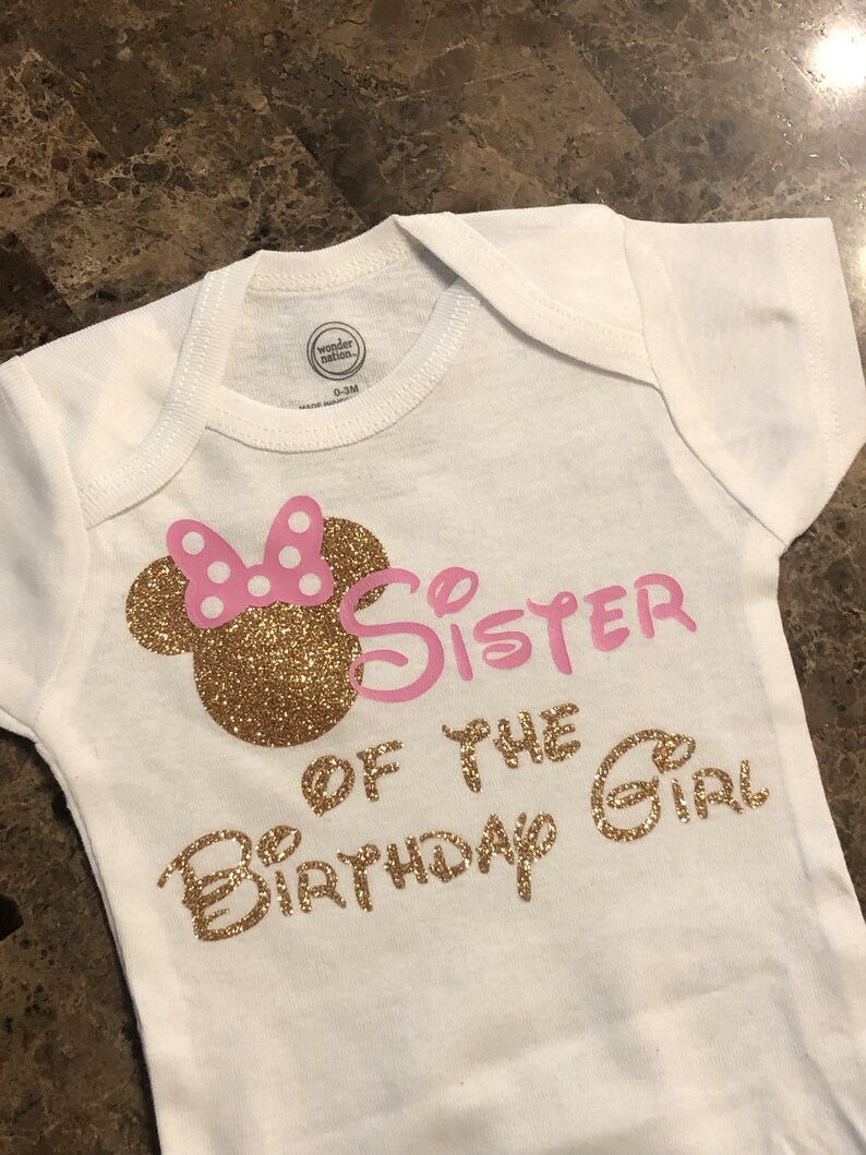 May include: White baby onesie with gold glitter lettering that says "Big Sister of the Birthday Girl". The onesie has a pink and gold glitter Minnie Mouse head on the left side.