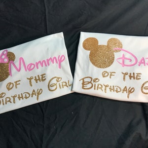 May include: Two white t-shirts with gold glitter Mickey Mouse ears and pink text. The text on the first shirt says "Mommy of the Birthday Girl". The text on the second shirt says "Dad of the Birthday Girl".