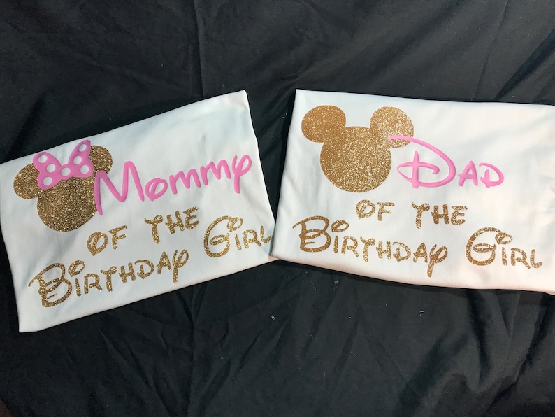 May include: Two white t-shirts with gold glitter Mickey Mouse ears and pink text. The text on the first shirt says "Mommy of the Birthday Girl". The text on the second shirt says "Dad of the Birthday Girl".