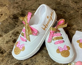May include: White canvas shoes with pink polka dot ribbon laces and gold glitter accents. The shoes have a gold glitter Minnie Mouse design on the toe and the name "HANNIGAH" written on the side.