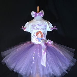May include: A white t-shirt with the text "Birthday Princess" and a cartoon graphic of a princess, paired with a layered lavender tulle skirt. The outfit includes a matching lavender bow and ribbon accents, displayed on a wooden mannequin.