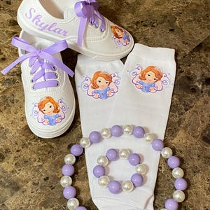 May include: White canvas sneakers with lavender laces and the name "Skylar" in script. Matching white socks and a beaded bracelet and necklace set in lavender and white. Each item features a princess graphic.