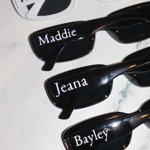K&ouml;nnte beinhalten: Sonnenbrillen mit personalisierten Namen. Das obere Paar ist wei&szlig; mit dem Namen "Kinde" auf dem Rahmen. Die anderen Paare sind schwarz mit den Namen "Maddie", "Jeana" und "Bayley" auf den Rahmen gedruckt.