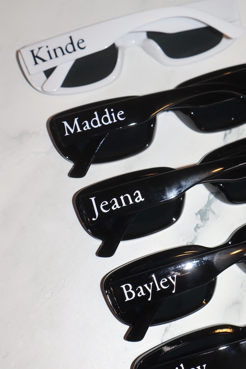 K&ouml;nnte beinhalten: Sonnenbrillen mit personalisierten Namen. Das obere Paar ist wei&szlig; mit dem Namen "Kinde" auf dem Rahmen. Die anderen Paare sind schwarz mit den Namen "Maddie", "Jeana" und "Bayley" auf den Rahmen gedruckt.
