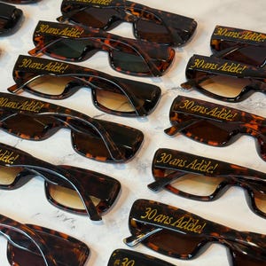 K&ouml;nnte beinhalten: Eine Sammlung von Sonnenbrillen im Schildpatt-Design mit Farbverlaufsgl&auml;sern. Jede Brille tr&auml;gt den goldenen Schriftzug "30 ans Ad&egrave;le!". Die Sonnenbrillen sind auf einer hellen Oberfl&auml;che angeordnet.