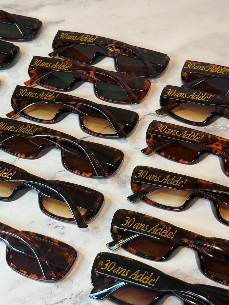 K&ouml;nnte beinhalten: Eine Sammlung von Sonnenbrillen im Schildpatt-Design mit Farbverlaufsgl&auml;sern. Jede Brille tr&auml;gt den goldenen Schriftzug "30 ans Ad&egrave;le!". Die Sonnenbrillen sind auf einer hellen Oberfl&auml;che angeordnet.
