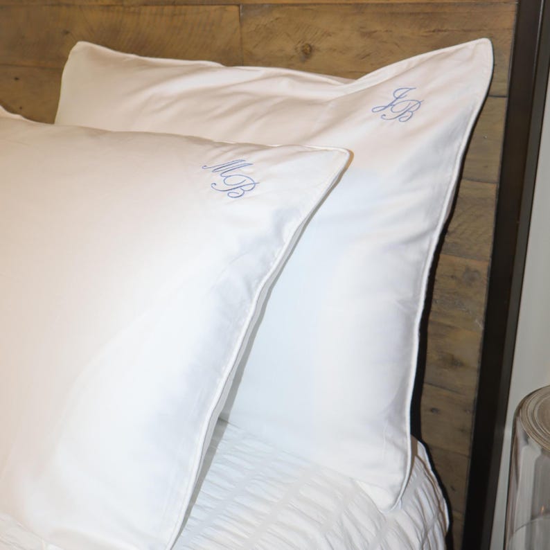Two white pillowcases, one in front of the other, with "MB" embroidered in light blue script on the front one and "JB" on the back one, against a wooden headboard.