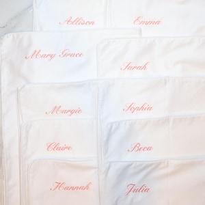 Eight white pillowcases are stacked, each embroidered with a different name in pink script: Allison, Emma, Mary Grace, Sarah, Margie, Sophia, Claire, Hannah, and Julia.