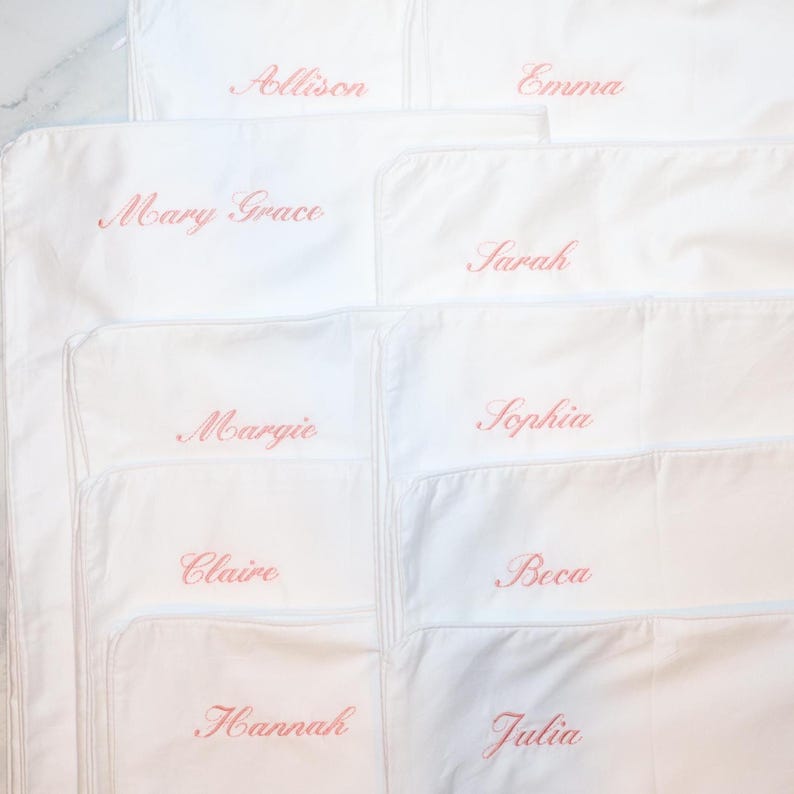 Eight white pillowcases are stacked, each embroidered with a different name in pink script: Allison, Emma, Mary Grace, Sarah, Margie, Sophia, Claire, Hannah, and Julia.