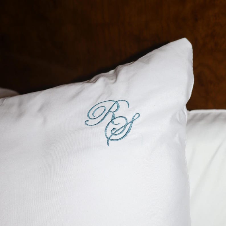 Puede incluir: Almohada blanca con iniciales azules bordadas en escritura cursiva. La almohada est&aacute; hecha de una tela suave y lisa. El fondo es de color marr&oacute;n oscuro.