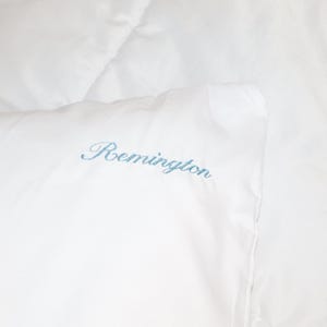 Puede incluir: Primer plano de ropa de cama blanca con el nombre "Remington" bordado en escritura azul claro. La tela parece suave y lisa. La imagen muestra una vista de cerca de la esquina de la ropa de cama.