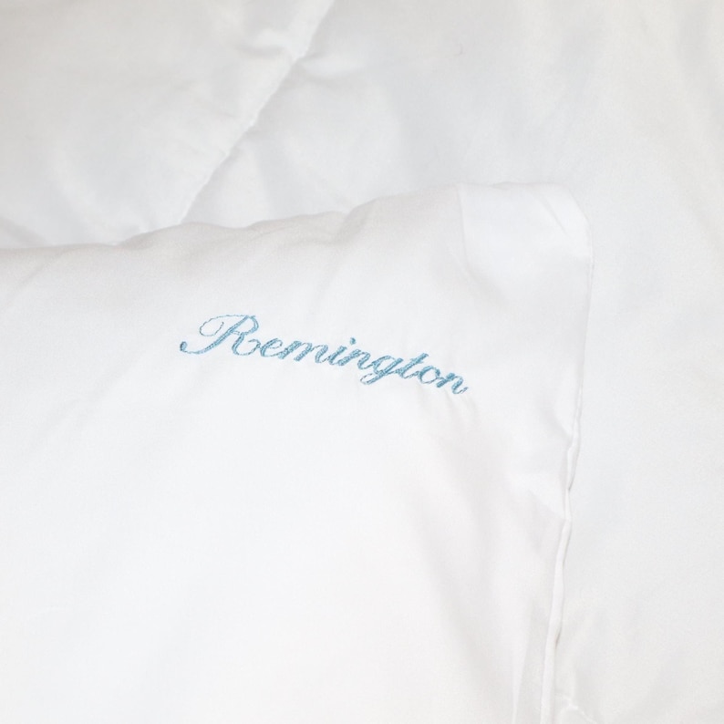 Puede incluir: Primer plano de ropa de cama blanca con el nombre "Remington" bordado en escritura azul claro. La tela parece suave y lisa. La imagen muestra una vista de cerca de la esquina de la ropa de cama.