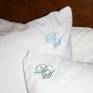 Puede incluir: Primer plano de almohadas blancas con iniciales bordadas. Una almohada tiene iniciales turquesas, mientras que otra tiene iniciales verdes. Las almohadas est&aacute;n sobre una cama, con un cabecero de madera al fondo.