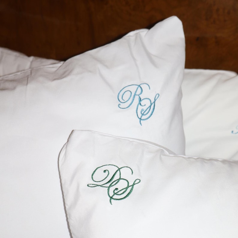 Puede incluir: Primer plano de almohadas blancas con iniciales bordadas. Una almohada tiene iniciales turquesas, mientras que otra tiene iniciales verdes. Las almohadas est&aacute;n sobre una cama, con un cabecero de madera al fondo.